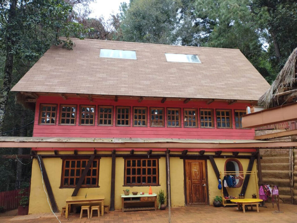 A two-story house with a sloped roof, yellow lower level, red upper level, multiple square windows, wooden door, benches, and potted plants in front, surrounded by trees.