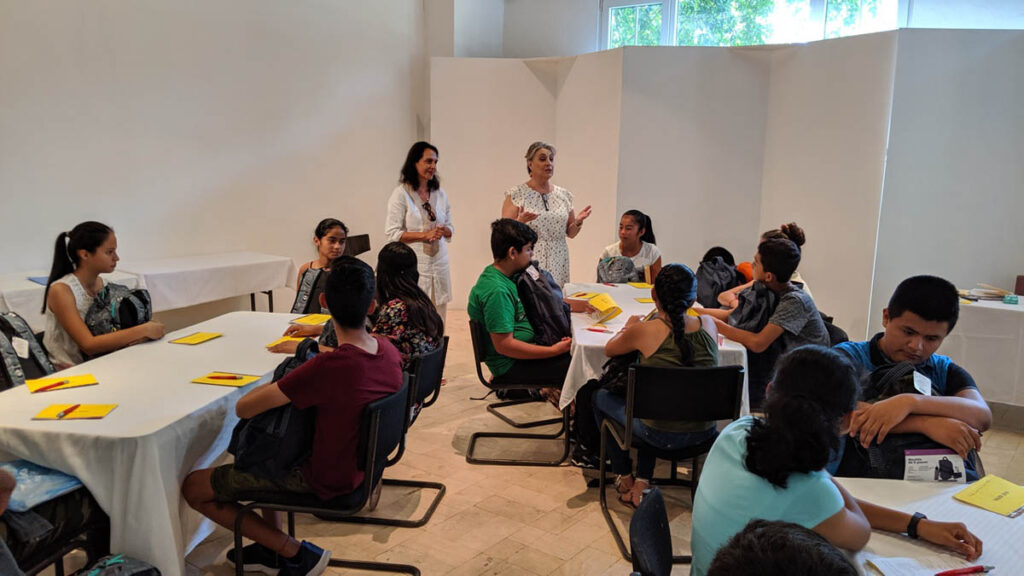 Two women stand and address a group of students seated at round tables with yellow papers in a classroom with white walls and natural light.