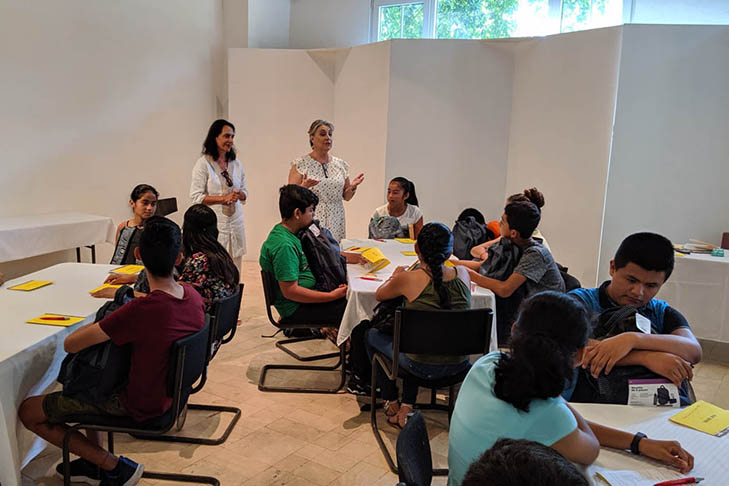 Two women stand and speak to a group of seated students at tables in a classroom, with papers and backpacks visible on the tables.
