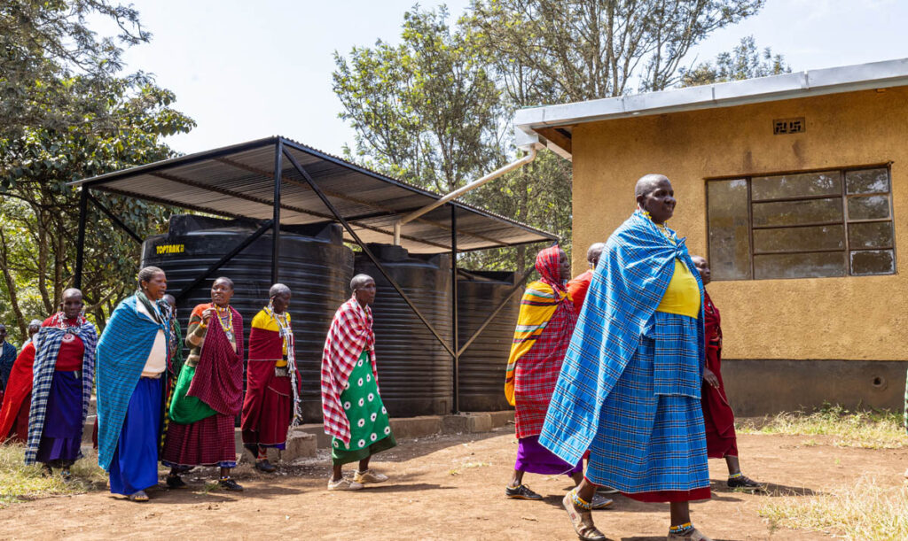 A group of people in colorful traditional clothing walk near a building and large water tanks outdoors on a sunny day.