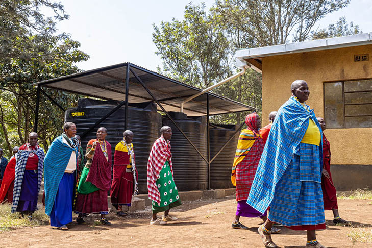 A group of people in colorful traditional clothing walk past large black water tanks and a building in an outdoor setting.