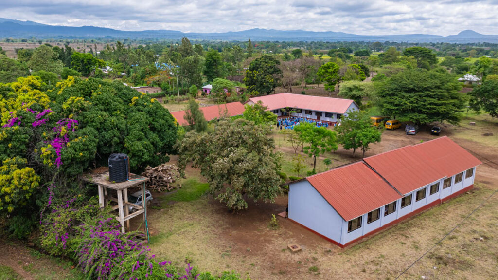 Aerial view of a rural school with red-roofed buildings, trees, a water tank on a tower, and open fields under a cloudy sky.