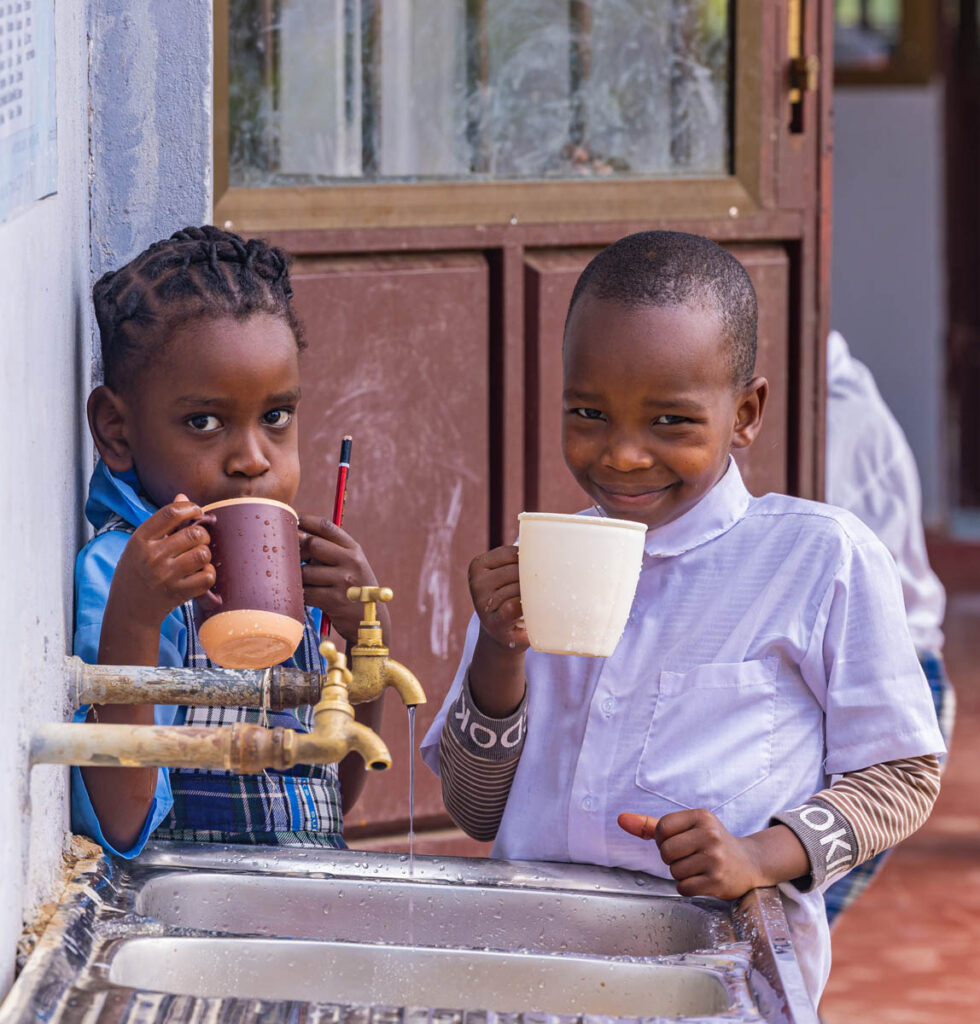Two children stand by a sink, holding mugs and drinking water, with faucets running and an open door in the background.