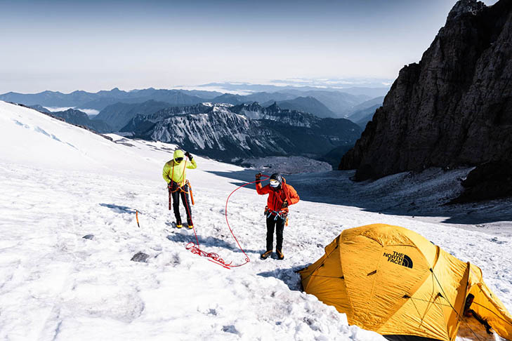 Two climbers in bright jackets handle ropes near a yellow tent on a snowy mountain slope, with rugged peaks and valleys visible in the background.