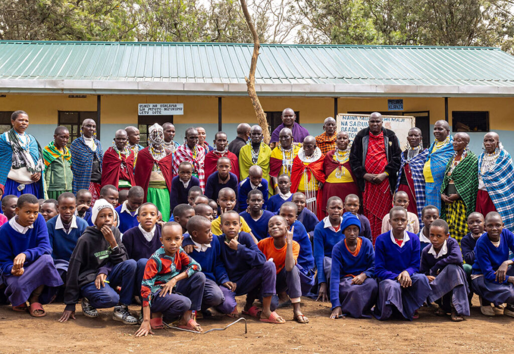 A group of school children in uniforms sit and kneel in front of a group of adults in colorful clothing, with a school building in the background.