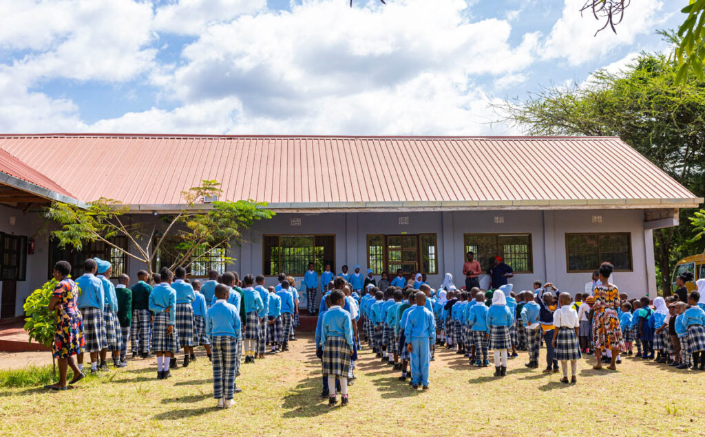 A large group of schoolchildren in blue uniforms stand in rows outside a school building during an outdoor assembly.