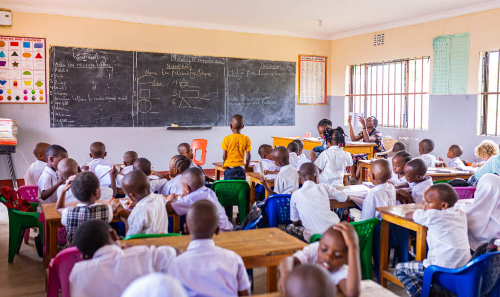 A classroom with young students seated at desks, working and writing, while a teacher and a child stand at the blackboard with geometric diagrams and notes written on it.