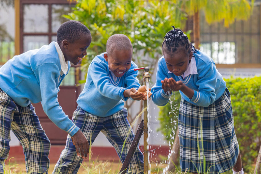 Three children in school uniforms happily gather around an outdoor water tap, catching and drinking water with their hands.