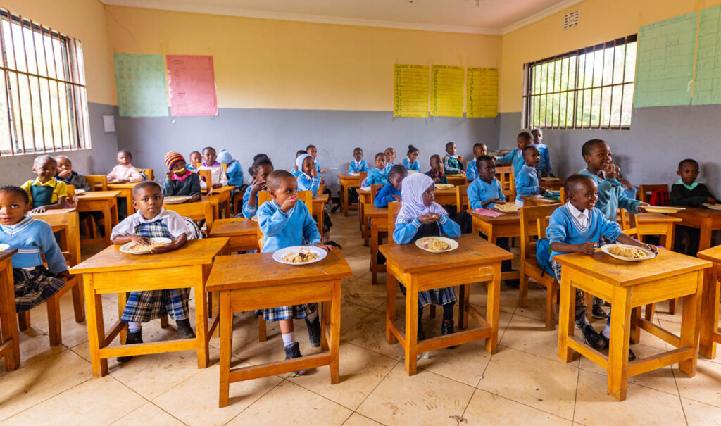 Children in blue uniforms sit at wooden desks in a classroom, eating meals from plates. The classroom has light-colored walls, large windows, and posters on the wall.