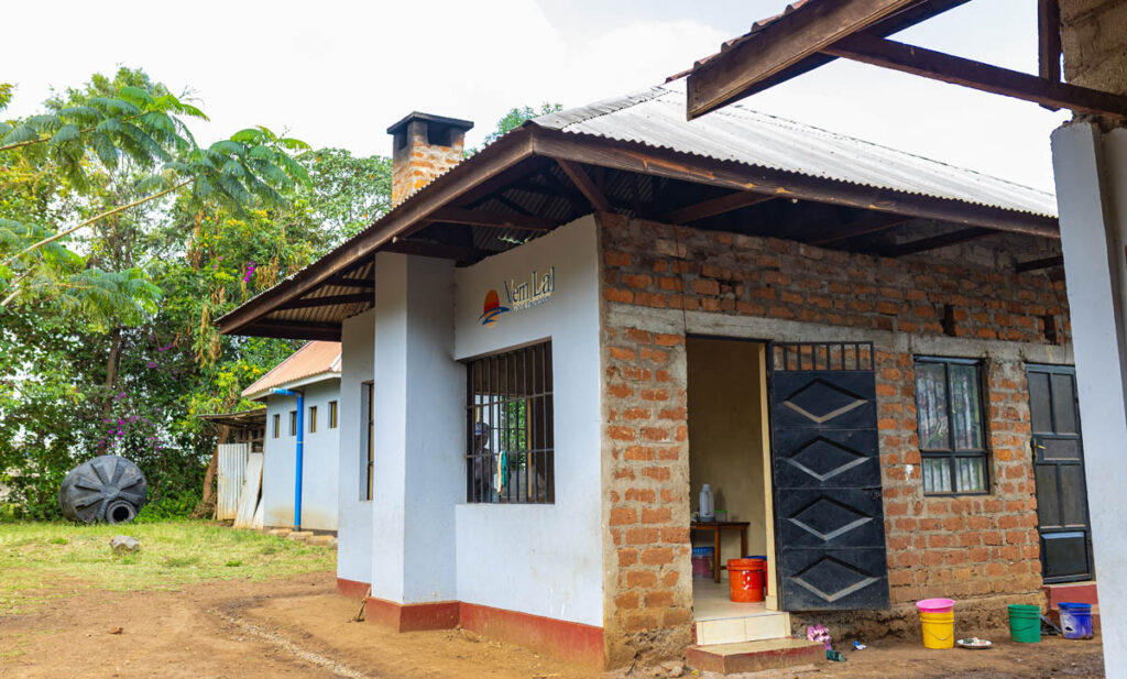 A brick building with a tin roof and open door stands next to a blue house; buckets and a black water tank are visible outside.