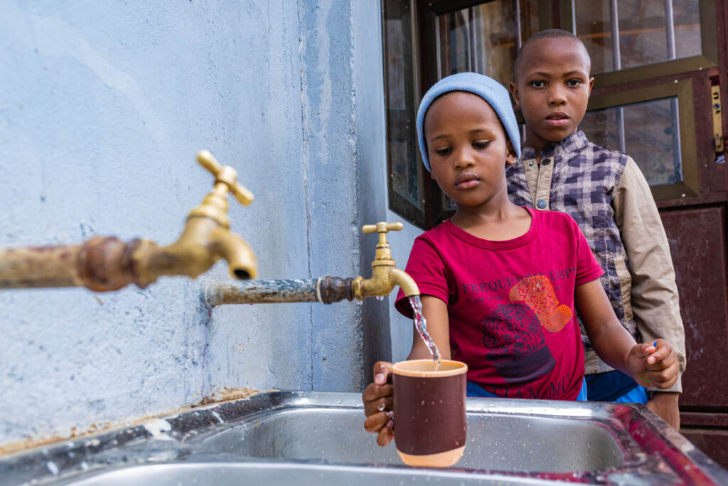 Two children stand by a sink as one fills a cup with water from a faucet attached to a wall.