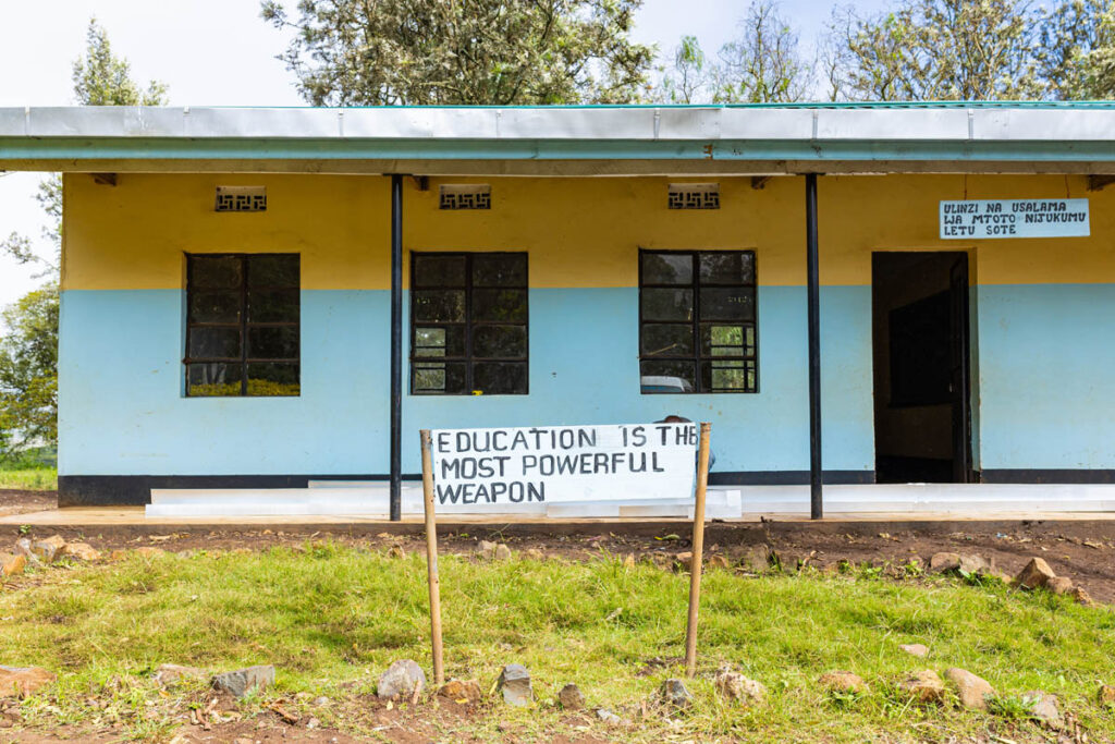 A single-story school building with blue and yellow walls; a sign in front reads, Education is the most powerful weapon.