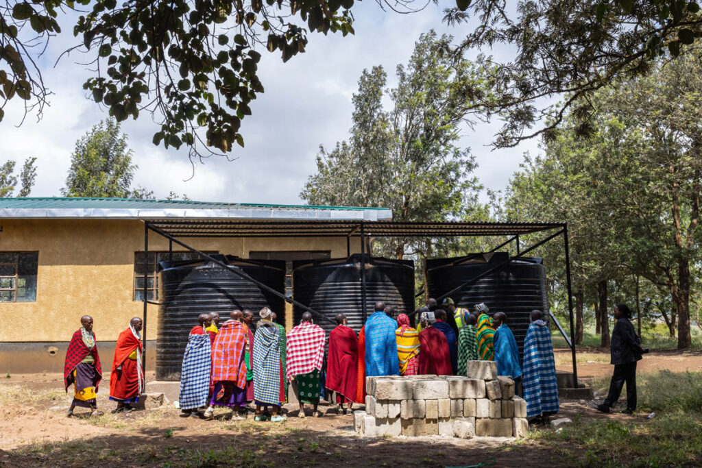 A group of people in colorful clothing gathers around large black water tanks outside a yellow building surrounded by trees.