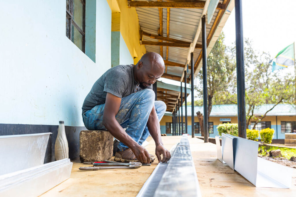 A man crouches on a concrete walkway, measuring and assembling metal gutters with various tools beside him, next to a building with blue walls and a corrugated metal roof.