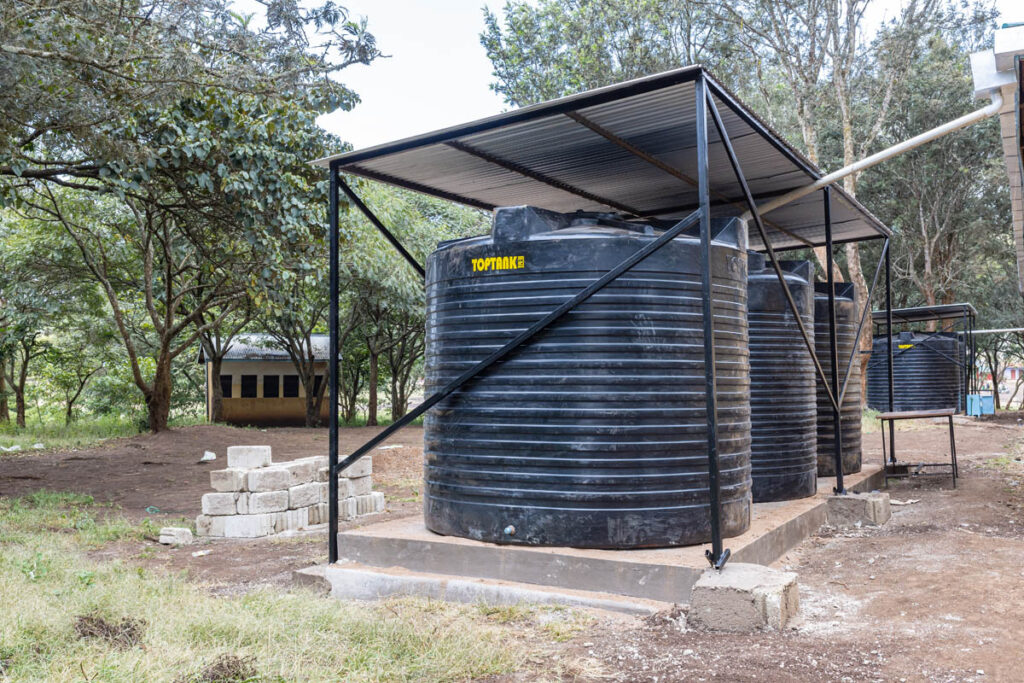 Two large black water storage tanks with a metal roof stand on a concrete platform outdoors, surrounded by trees and some concrete blocks.