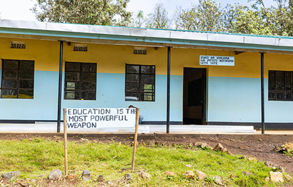 A single-story school building with blue and yellow walls has a sign in front that reads, EDUCATION IS THE MOST POWERFUL WEAPON.