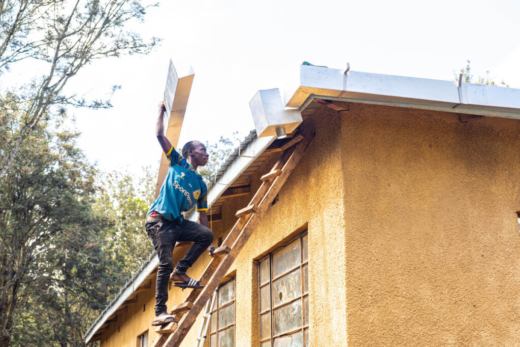 A man climbs a wooden ladder to the roof of a yellow building while carrying metal sheets, with trees visible in the background.