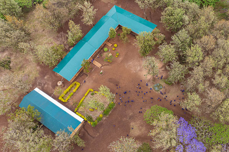 Aerial view of two buildings with green roofs surrounded by trees, dirt pathways, and groups of people scattered around the open grounds.