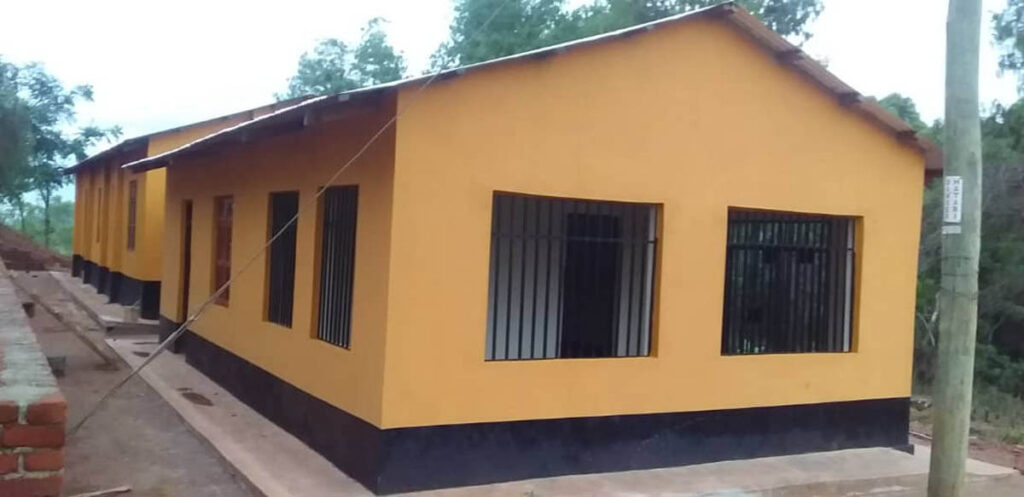 A single-story yellow building with barred windows and multiple doorways, set on a concrete foundation with trees in the background.