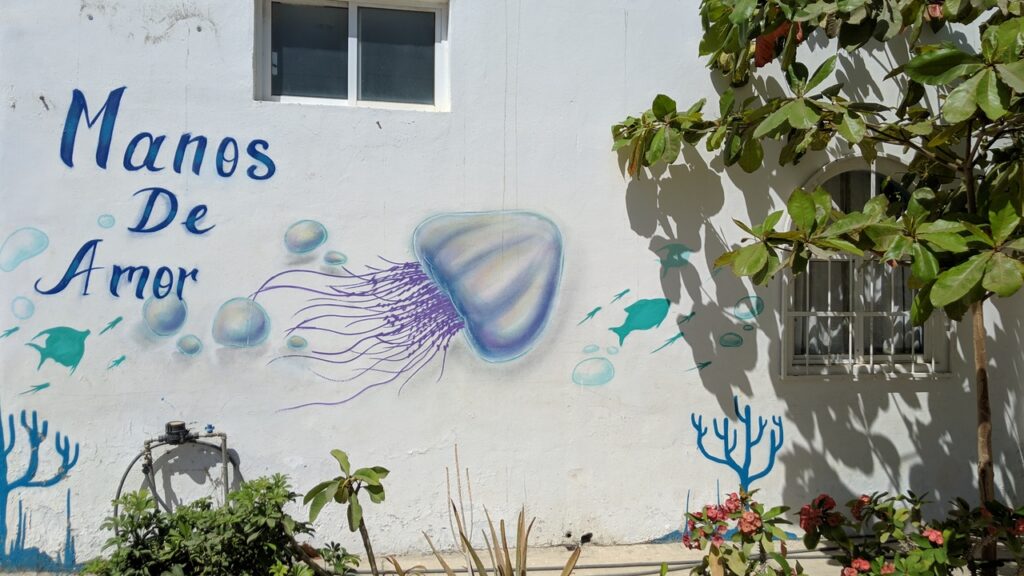 A mural on a white wall features a large jellyfish, small fish, coral, and the words Manos De Amor in blue, with plants and a window nearby.