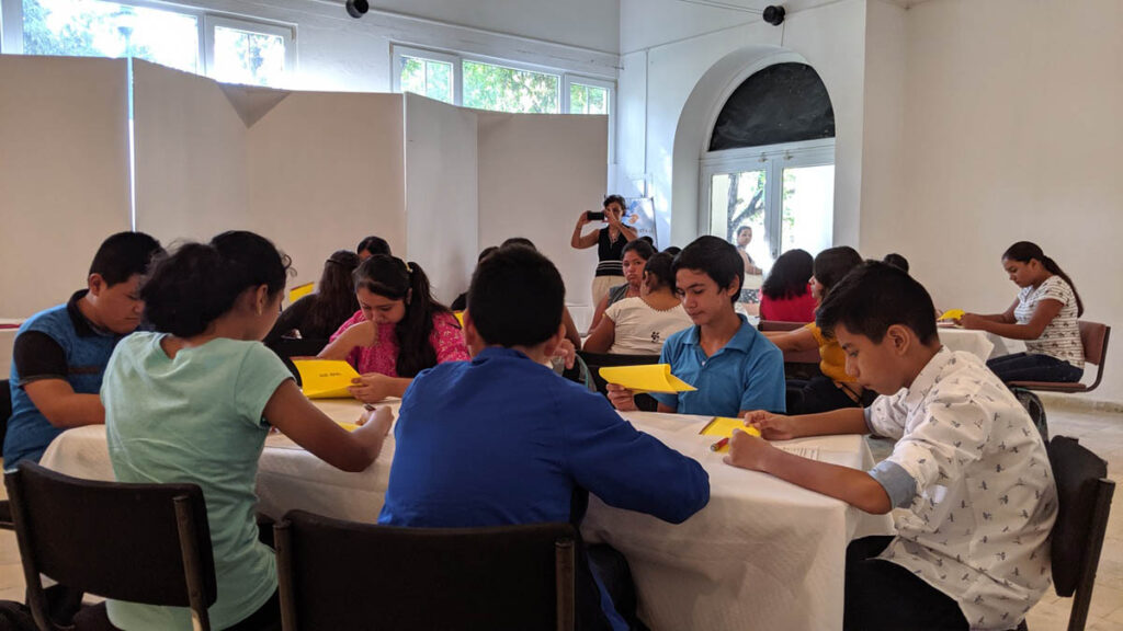 A group of students sit around tables in a classroom, reading or writing on yellow papers, while an adult stands and takes a photo in the background.