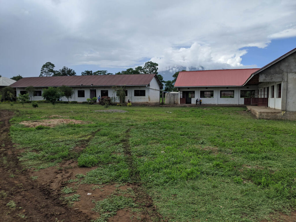 Three single-story buildings with red roofs stand around a grassy yard under a cloudy sky; a few people are visible near one building.