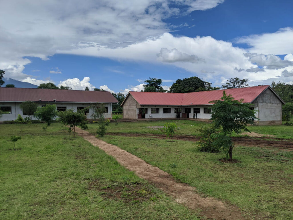 Two single-story buildings with red roofs are separated by a grassy yard and a dirt pathway under a partly cloudy sky.