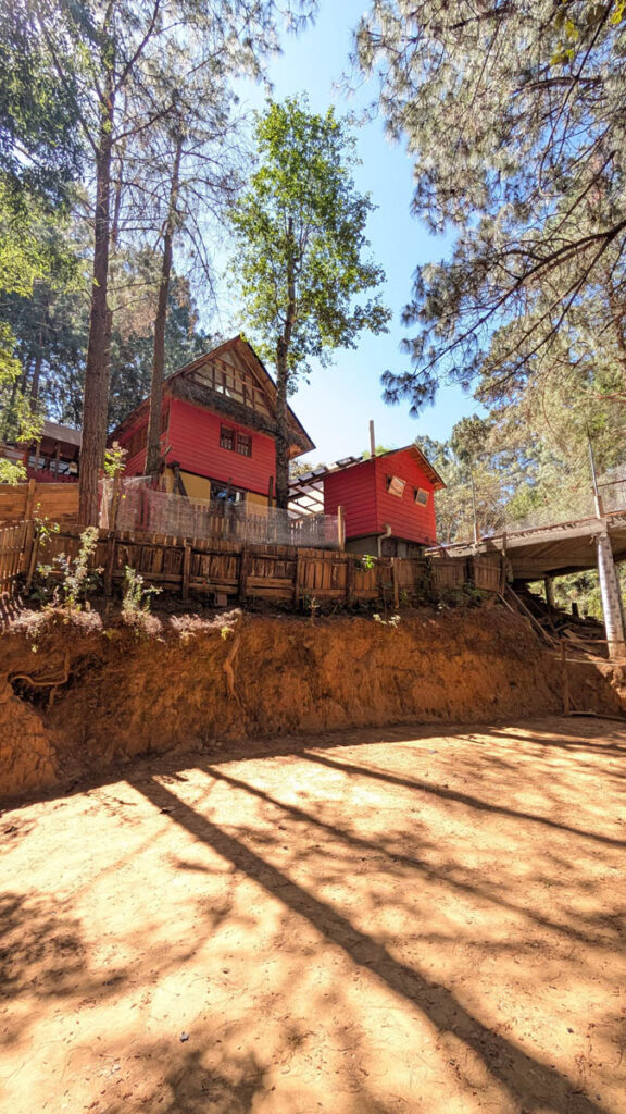 Two red wooden houses sit on a dirt slope surrounded by tall pine trees, with a wooden fence and a bridge on the right side under a clear blue sky.