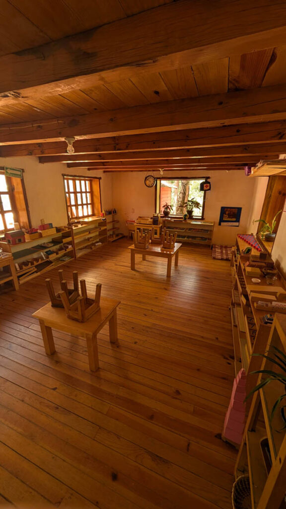 A wooden classroom with tables, chairs stacked upside down, shelves with educational materials, large windows, and exposed wooden beams on the ceiling.