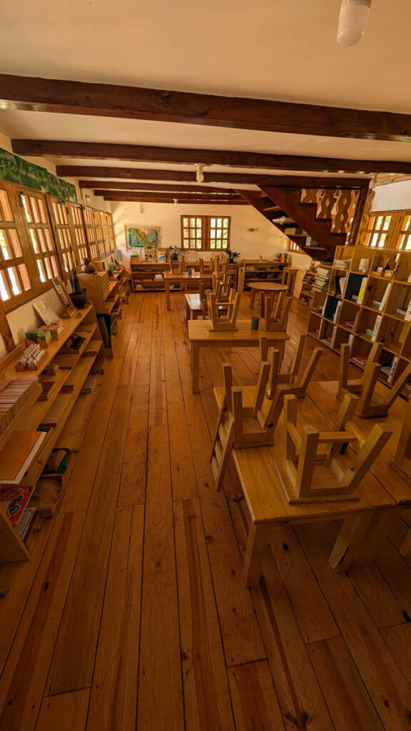 Classroom with wooden floors and furniture, chairs placed upside down on tables, bookshelves along the walls, and large windows letting in natural light.