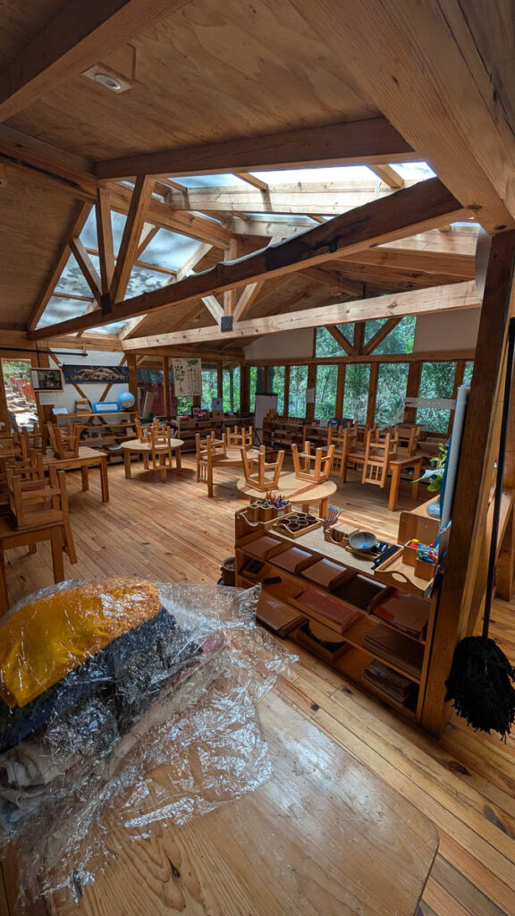 Wooden chairs and tables are neatly arranged in a spacious, sunlit room with exposed beams and large windows, with some folded blankets and bags in the foreground.