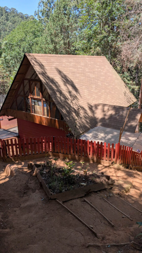 A-frame wooden cabin with large windows, surrounded by trees, a red picket fence, and a small garden bed in the foreground.