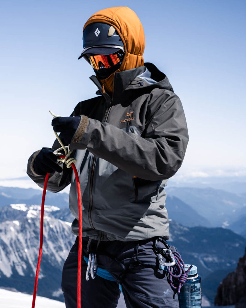 Person in mountaineering gear ties a red rope. Snowy mountains and blue sky are visible in the background.
