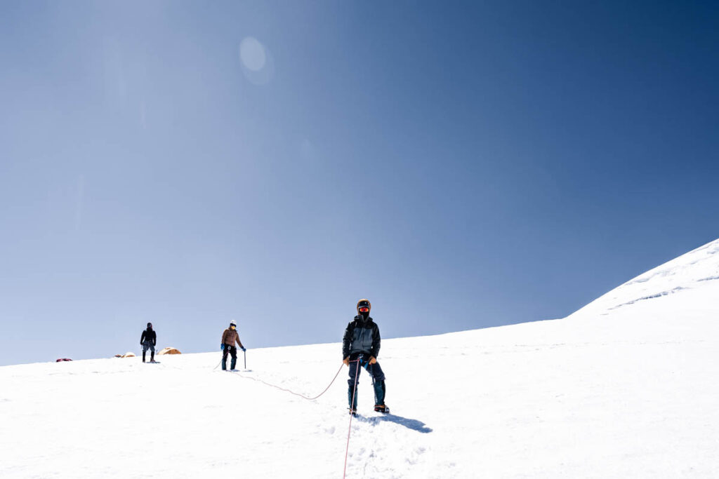 Three climbers roped together ascend a snowy slope under a clear blue sky, with tents visible in the distance.