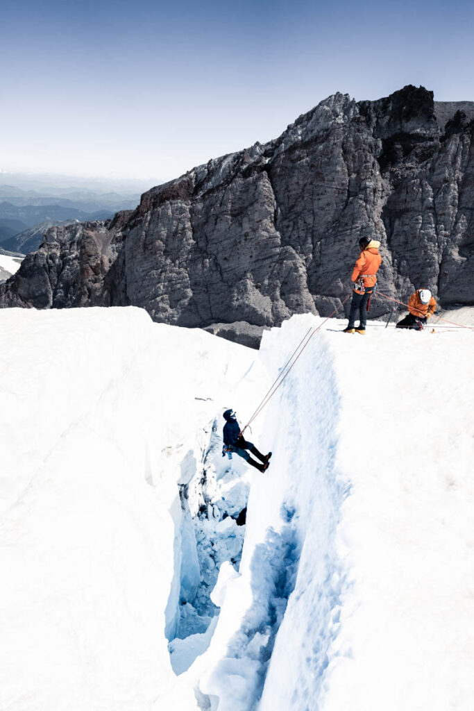 Three climbers in orange jackets are on a snowy mountain; one descends into a crevasse using ropes while two others stand at the edge. Rocky cliffs are visible in the background.