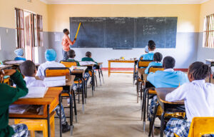 A teacher stands at the blackboard using a large ruler to point at text while students in uniforms sit at desks facing the front of a classroom.