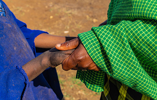 Two people are shaking hands outdoors; one wears a blue garment, and the other wears a green, checkered garment.