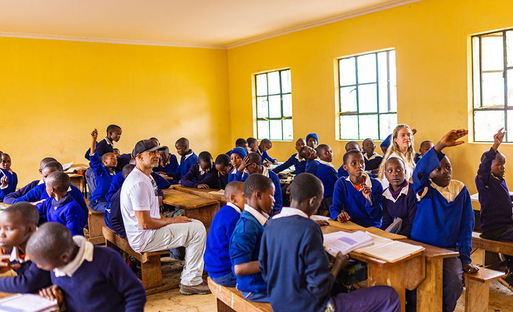 Students in blue uniforms sit at wooden desks in a brightly lit classroom with yellow walls, some raising hands, while two adults observe among them.