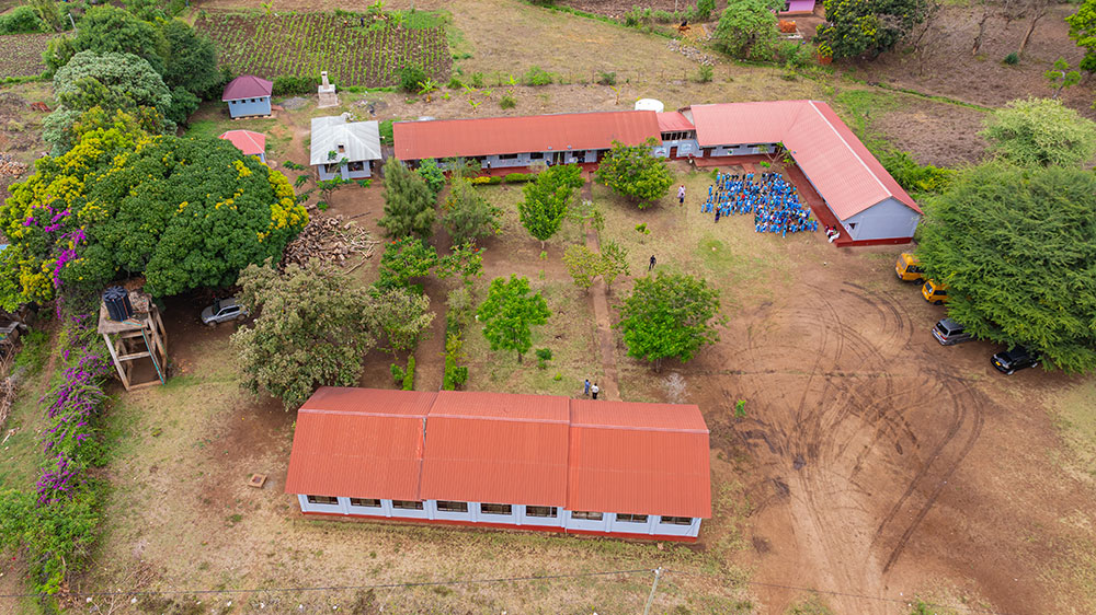 Aerial view of a rural school compound with red-roofed buildings, a group of people gathered outside, trees, and surrounding farmland.