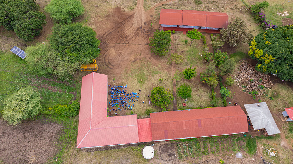 Aerial view of a rural school compound with red-roofed buildings, a group of students in blue uniforms, a yellow school bus, trees, and solar panels.