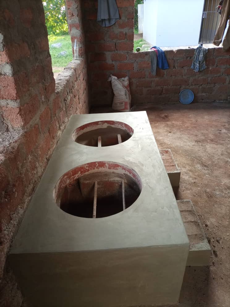 A newly constructed brick and cement stove with two round burner openings is situated in a partially built, open-walled room with unfinished brick walls and a cement floor.