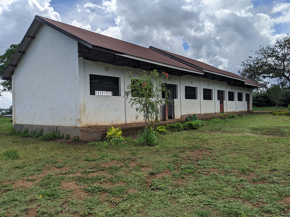 A single-story white building with a metal roof, barred windows, and a grassy area in front under a cloudy sky.