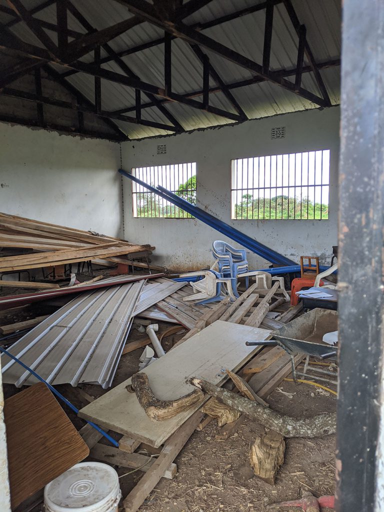 A cluttered room with scattered wooden boards, plastic chairs, and various debris piled on a dirt floor under a metal roof.