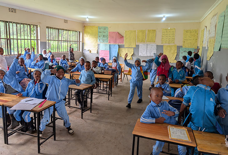 A classroom filled with students in blue uniforms, some seated and some standing, smiling and waving at the camera. Educational posters are displayed on the walls.