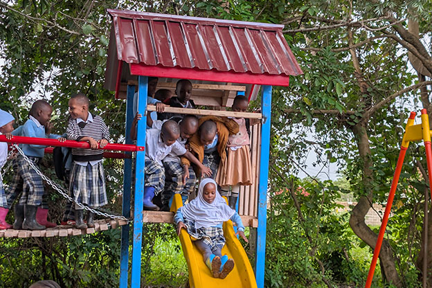 Children in school uniforms play on a playground structure; one girl is sliding down a yellow slide while others watch and wait above. Trees and greenery surround the area.
