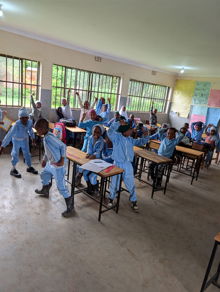 A classroom filled with students in blue uniforms, some standing and some sitting at desks, with large windows and educational posters on the walls.