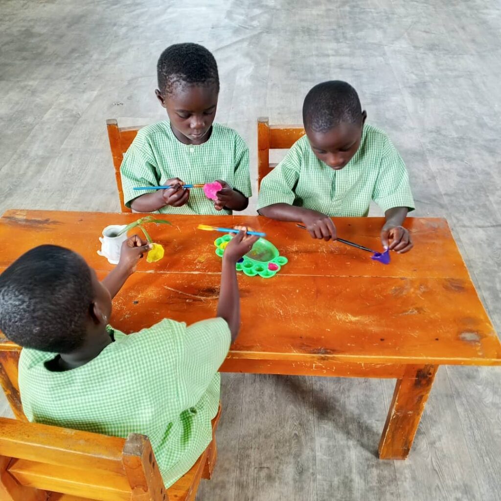 Three children in green uniforms sit at a wooden table, painting with brushes and colorful paints on a palette in a classroom setting.