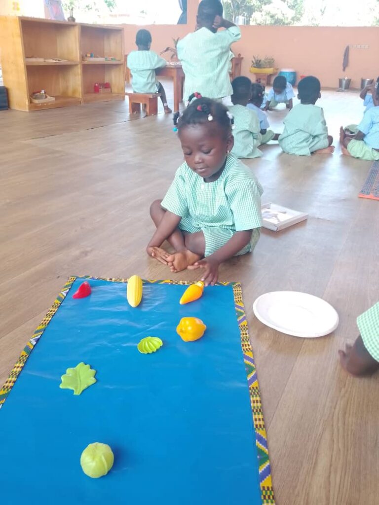 A young child in a green uniform sits on a wooden floor, arranging plastic fruits and vegetables on a blue mat, with other children visible in the background.