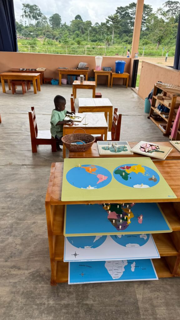 A child sits alone at a table in a spacious classroom. In the foreground, educational materials, including world maps, are displayed on a wooden shelf.