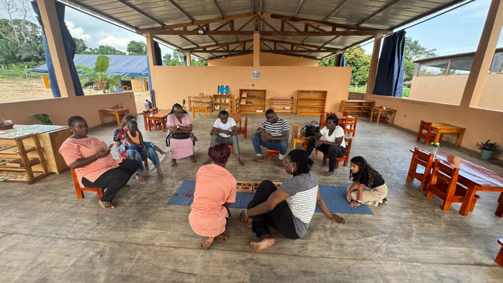 A group of people sit in a circle on mats and chairs in a covered open-air space, engaged in a group activity or discussion.
